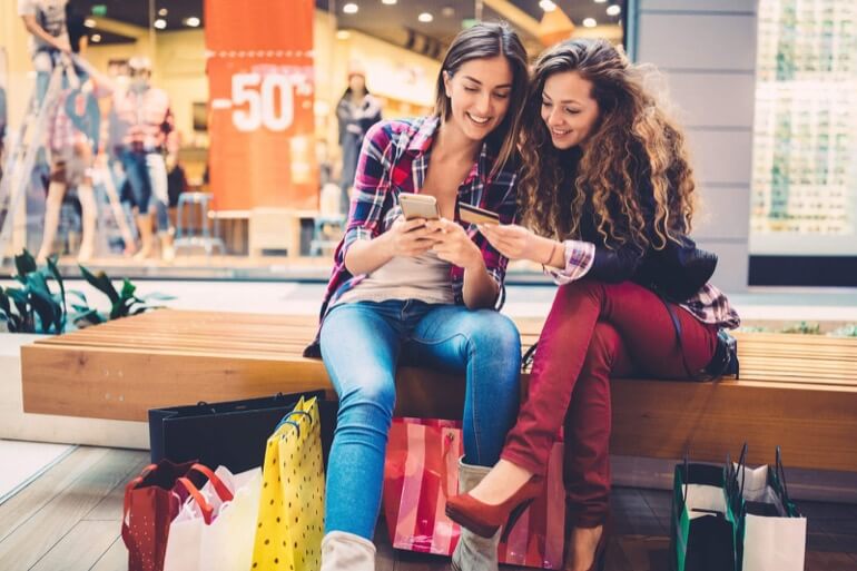 duas mulheres no shopping com várias sacolas e vendo celular